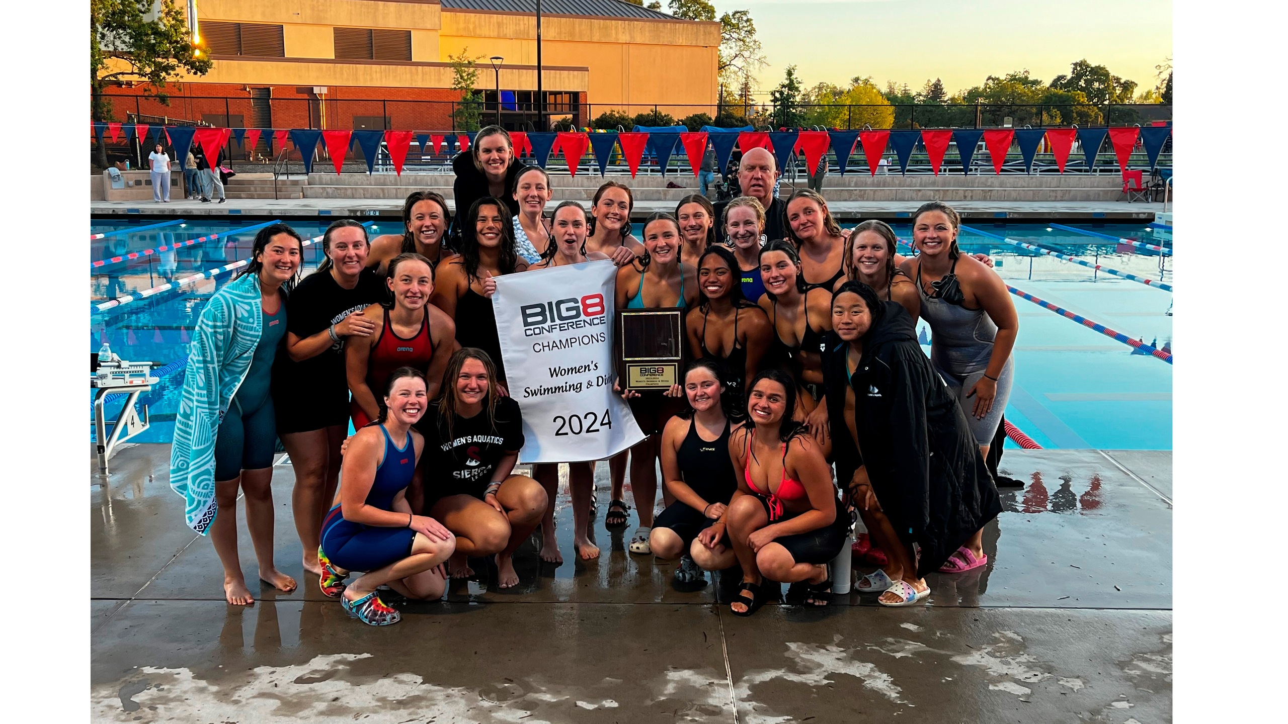 Women's swim dive pose with 2024 Big 8 championship banner and trophy in front of pool.